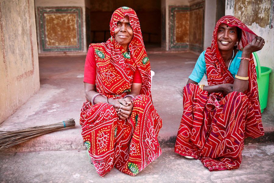 Two Indian women in vibrant red traditional Rajasthani saris sitting together smiling in a courtyard.