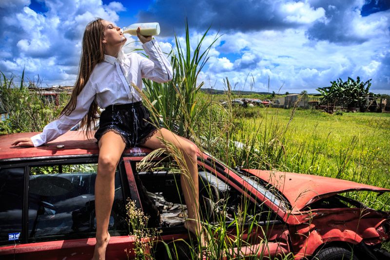 Young woman in white shirt sitting on red vintage car in rural field.