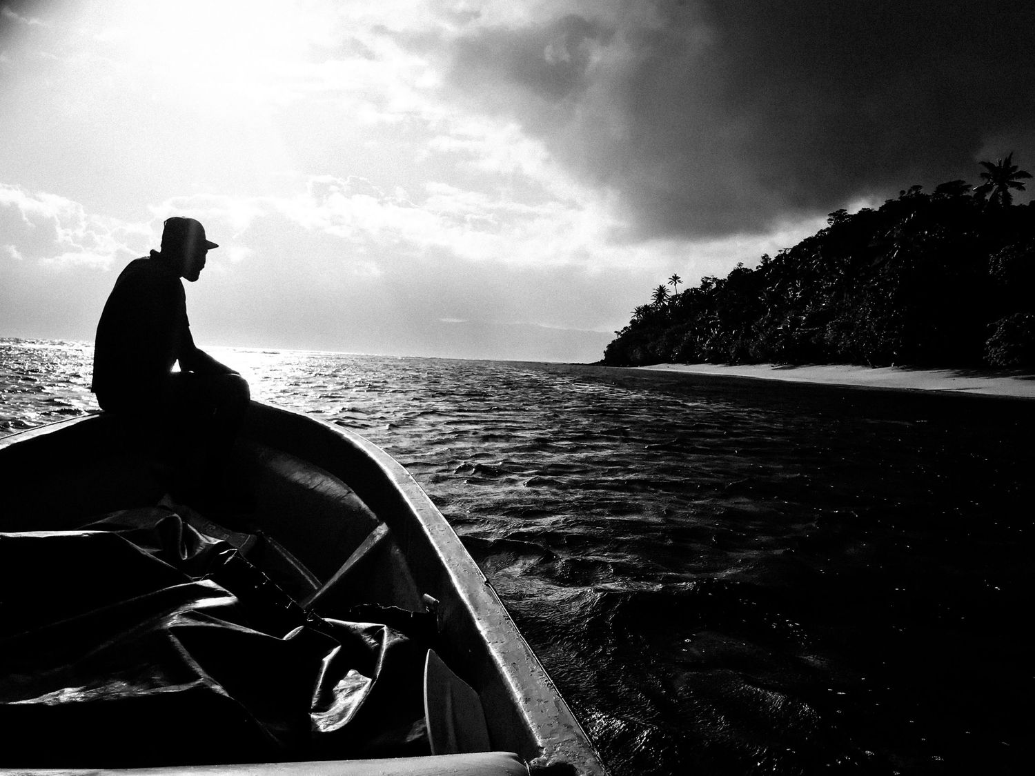 Black and white silhouette of a man sitting on a boat traveling along a tropical coastline at sunset.
