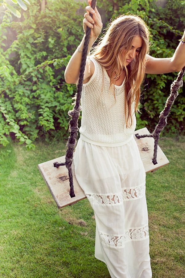 Woman in white crochet top and lace maxi skirt standing on a wooden swing in a lush green garden.