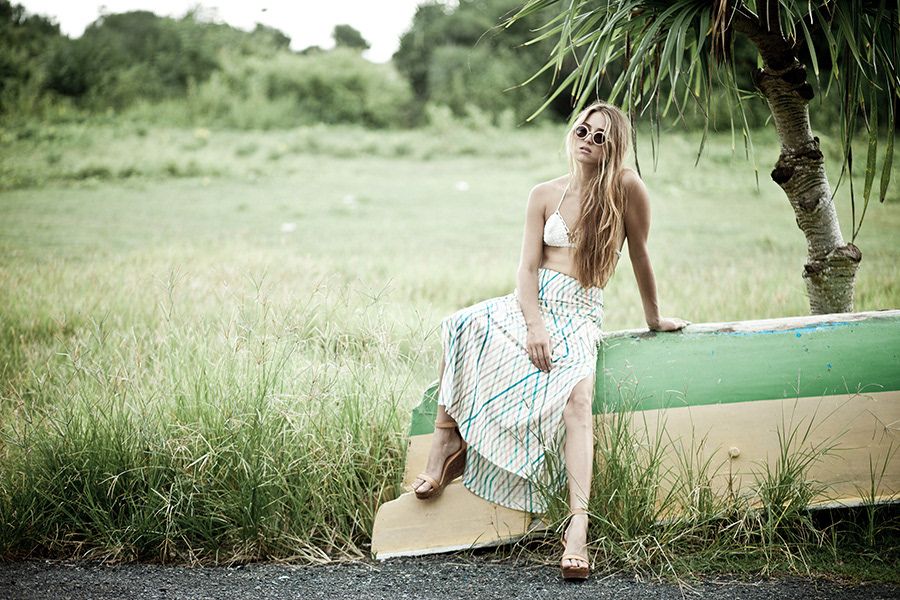 Blonde woman in white bikini top and maxi skirt sitting on old boat outdoors.