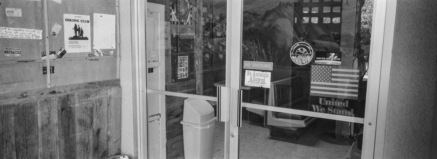 Black and white panoramic photo of a small commercial building entrance with glass doors.