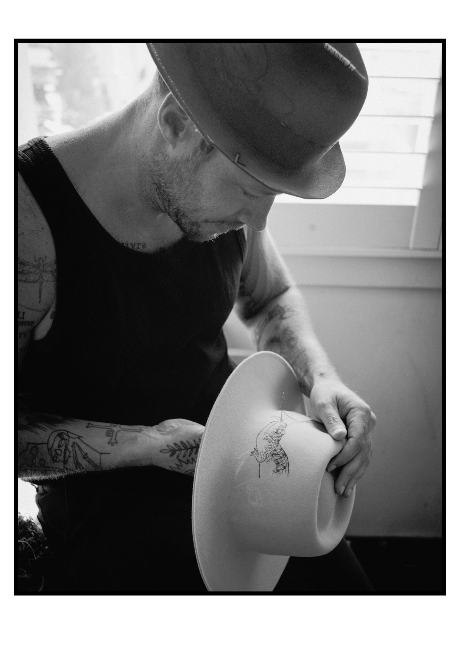 Tattooed hat maker signing a white wide-brim felt hat in black and white.