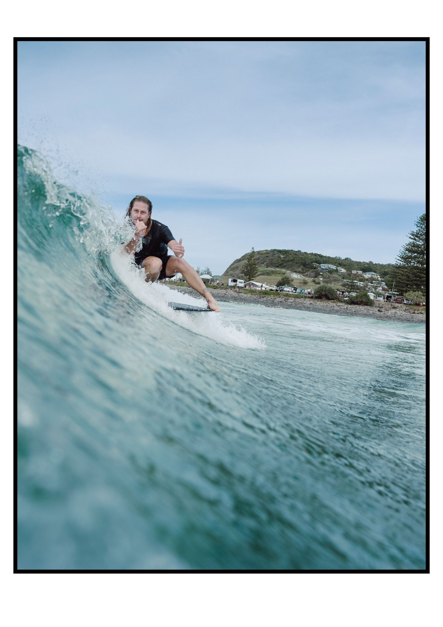 Surfer riding a turquoise wave near a coastal town on a cloudy day.