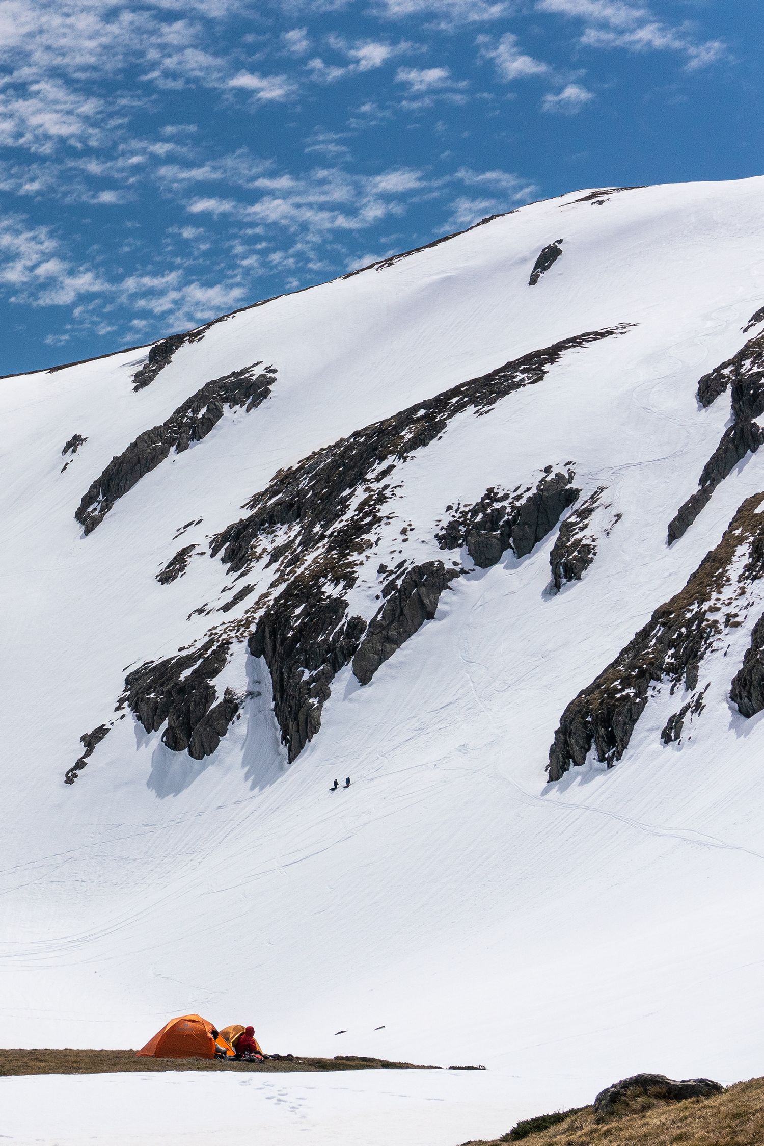 Orange camping tent at base of snow-covered mountain with climbers visible on slope.
