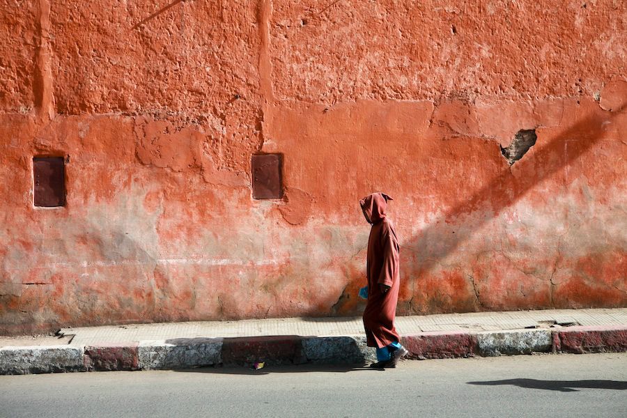 A person in a red hooded robe walks along a weathered terracotta red wall in Marrakech, Morocco.