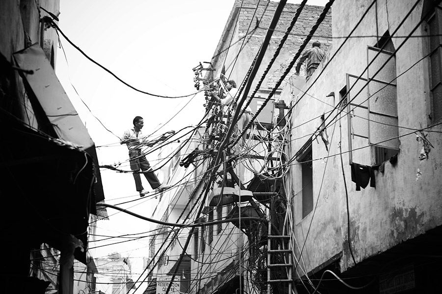 Black and white photo of a man balancing on electrical wires between buildings in a crowded urban neighborhood.