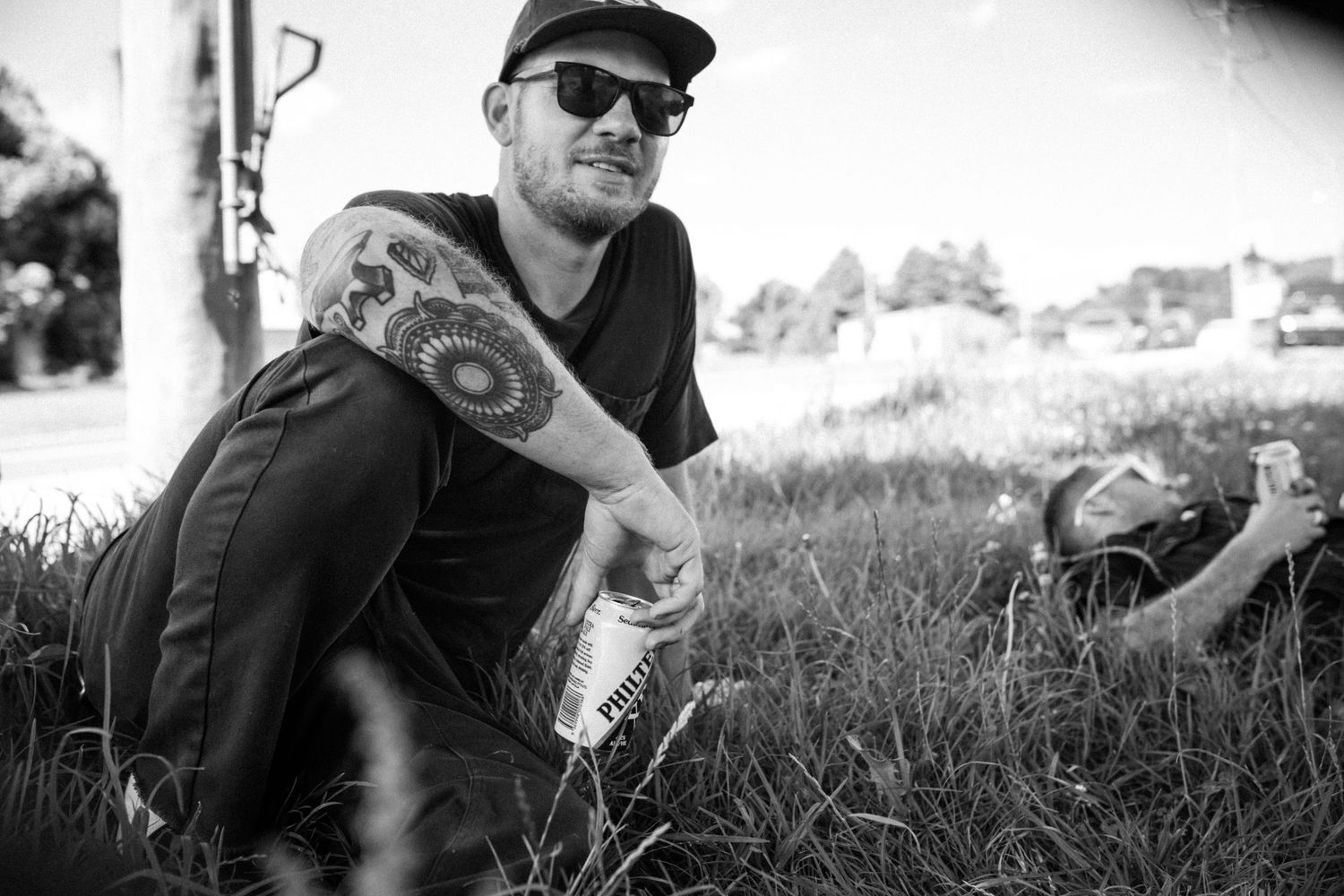 Black and white photo of a tattooed man in sunglasses sitting in grass holding a can.