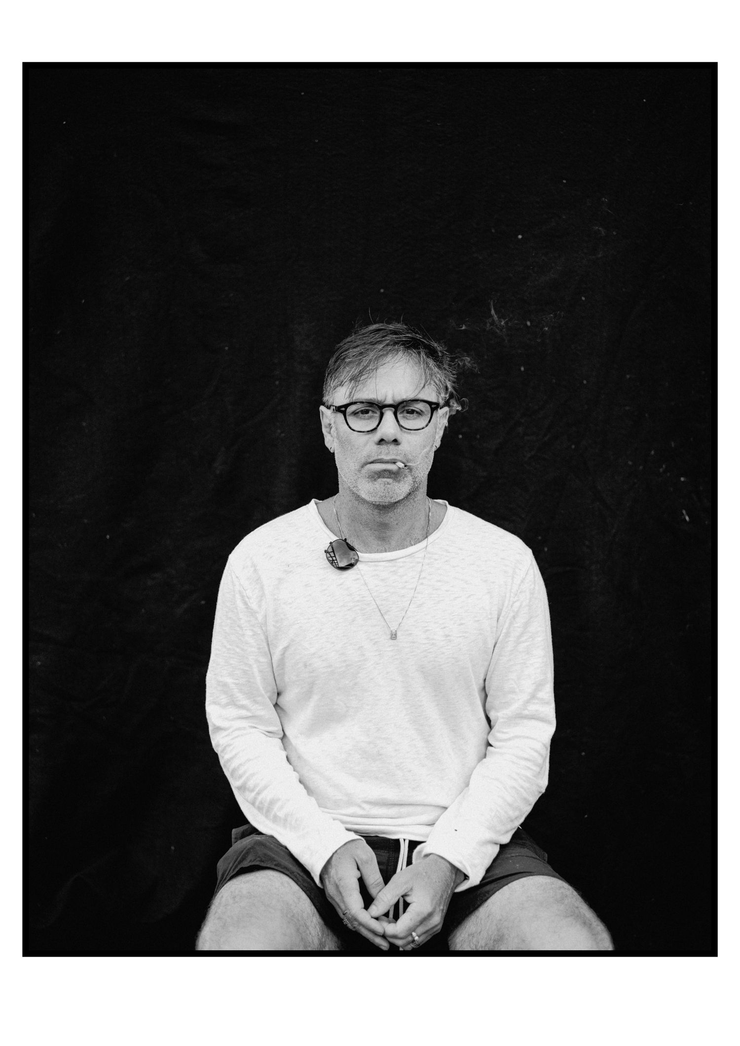 Black and white portrait of man with glasses in white sweatshirt seated against dark background.