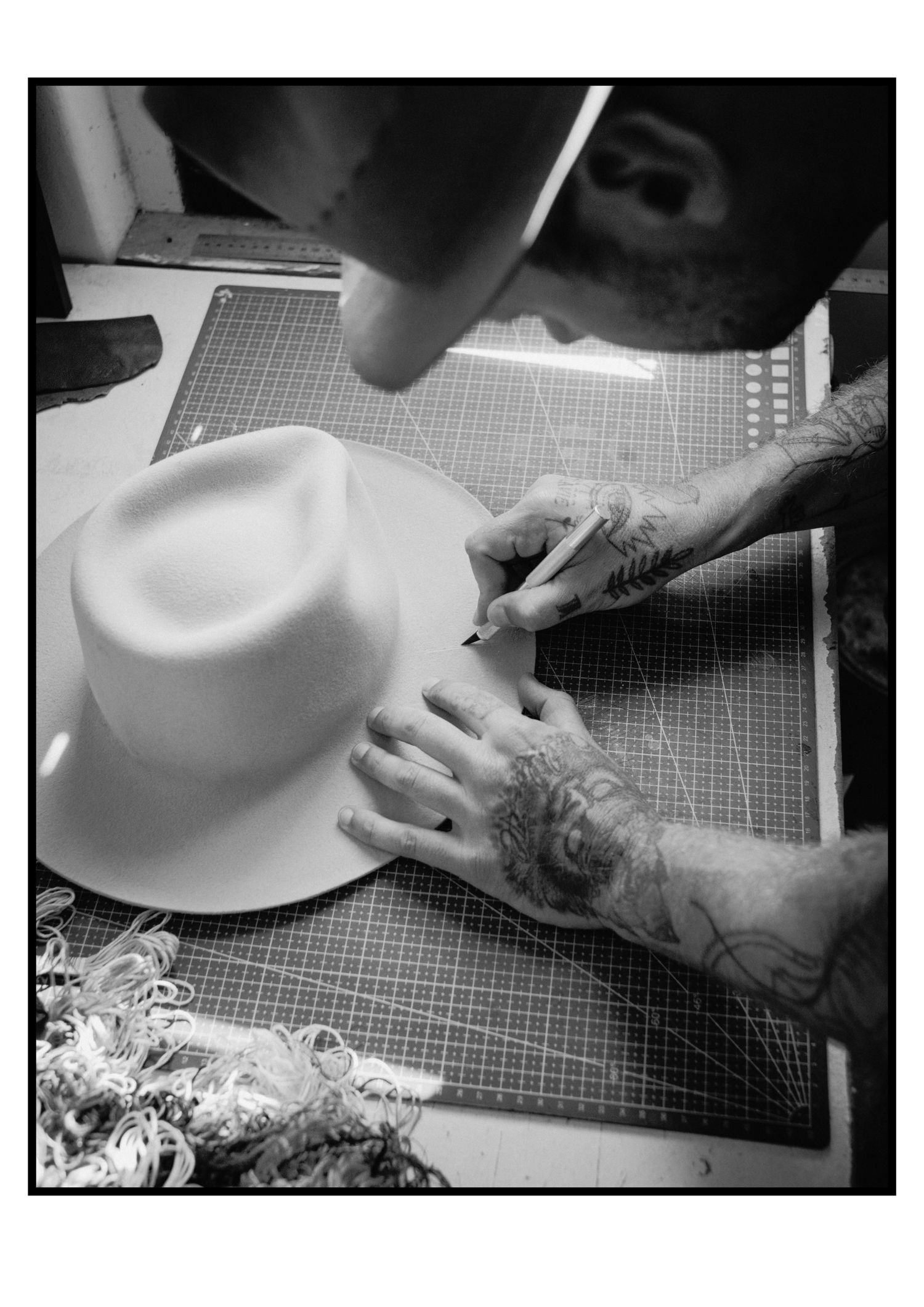 Tattooed hands drawing custom design on white wide-brim felt hat on cutting mat.