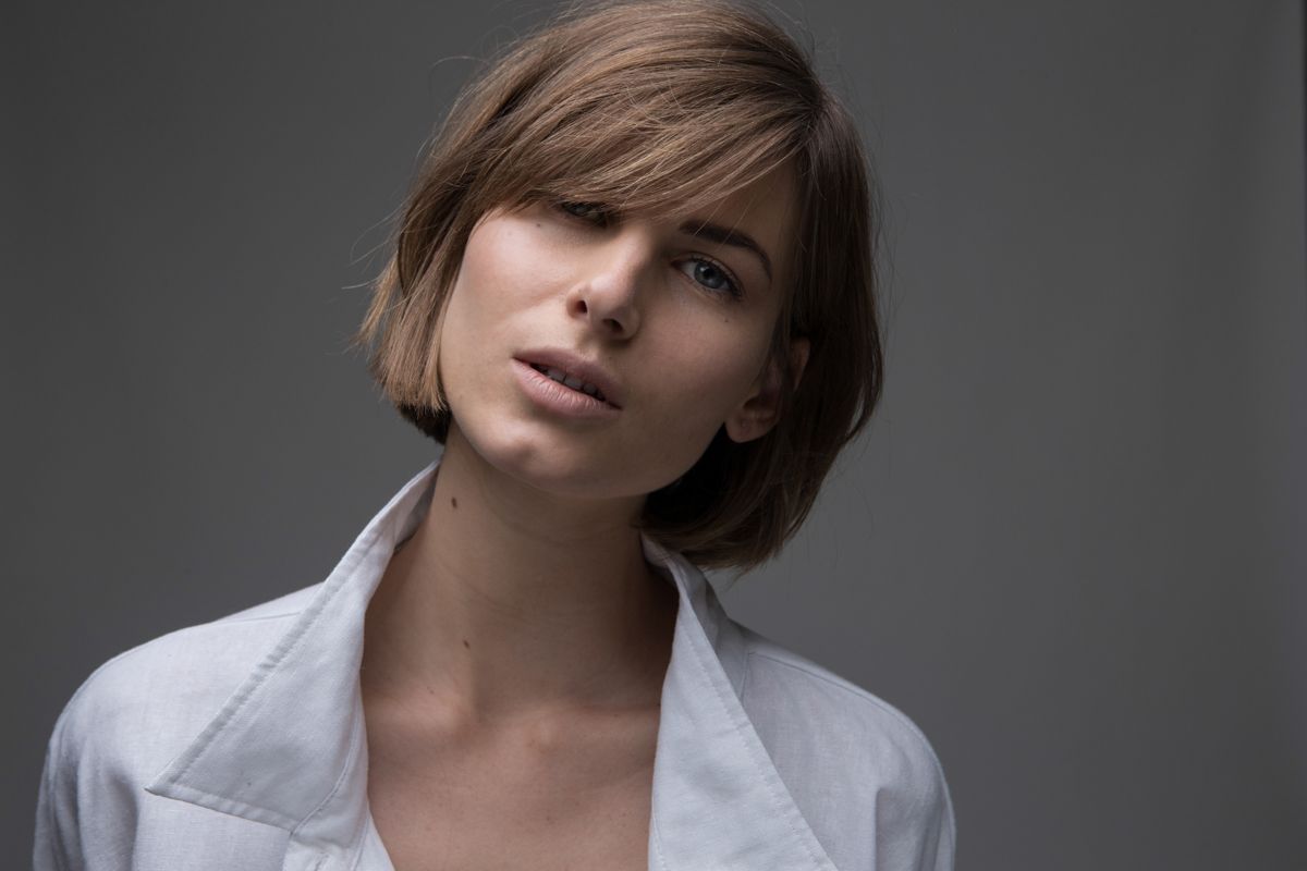 A young woman with a short bob haircut wearing a white shirt poses against a gray background.