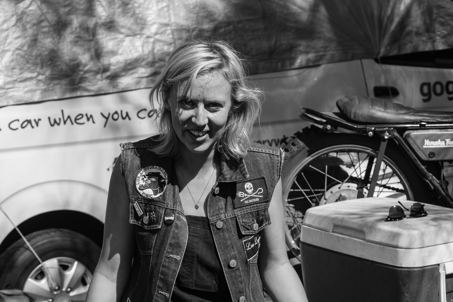 Smiling blonde woman in denim vest standing near a motorcycle and van in black and white photo.