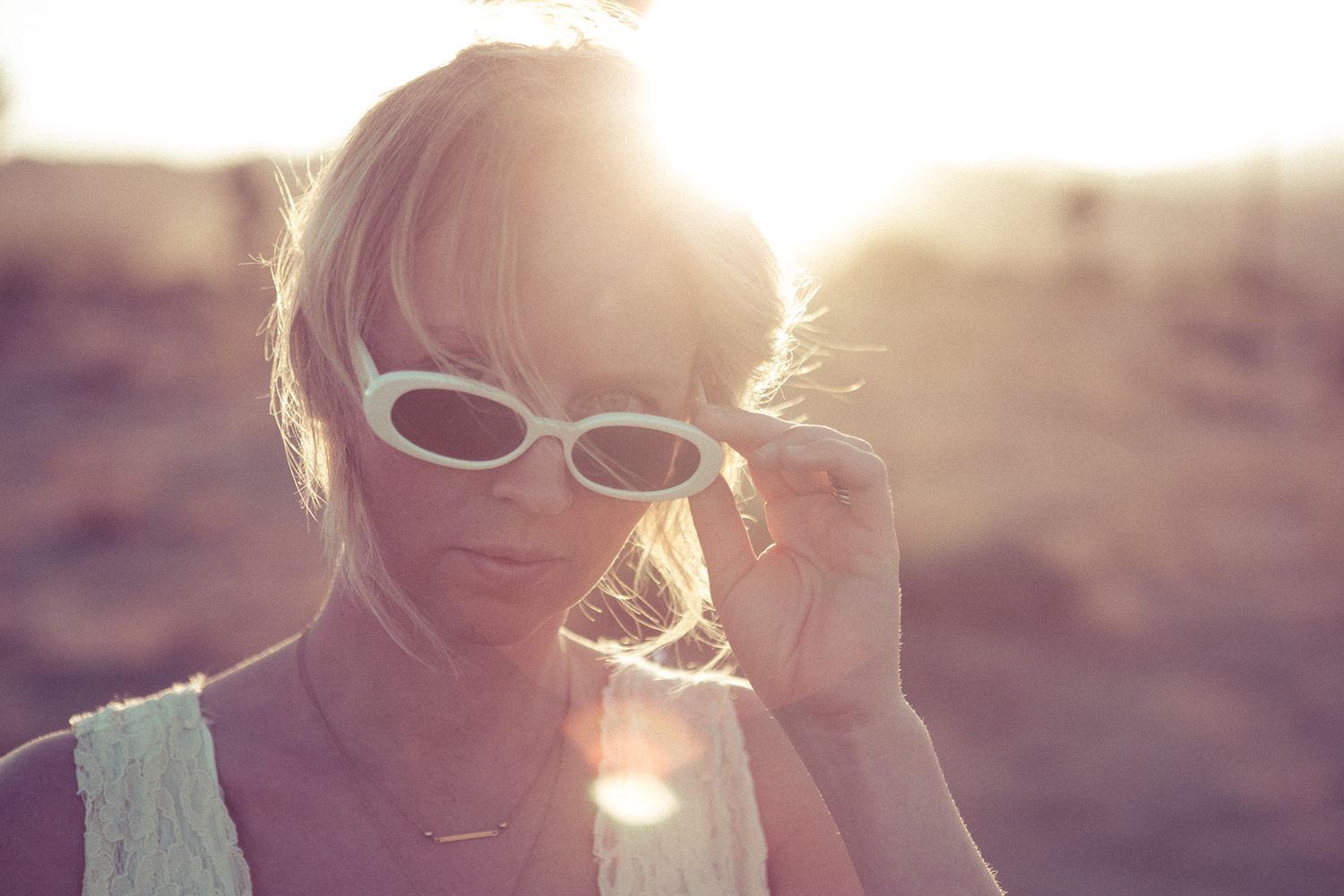 Blonde woman in white sunglasses and top posing outdoors in bright golden sunlight.
