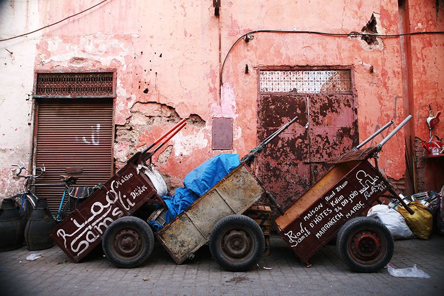 Rustic wooden market carts with handwritten text rest against a weathered pink wall in Marrakech.