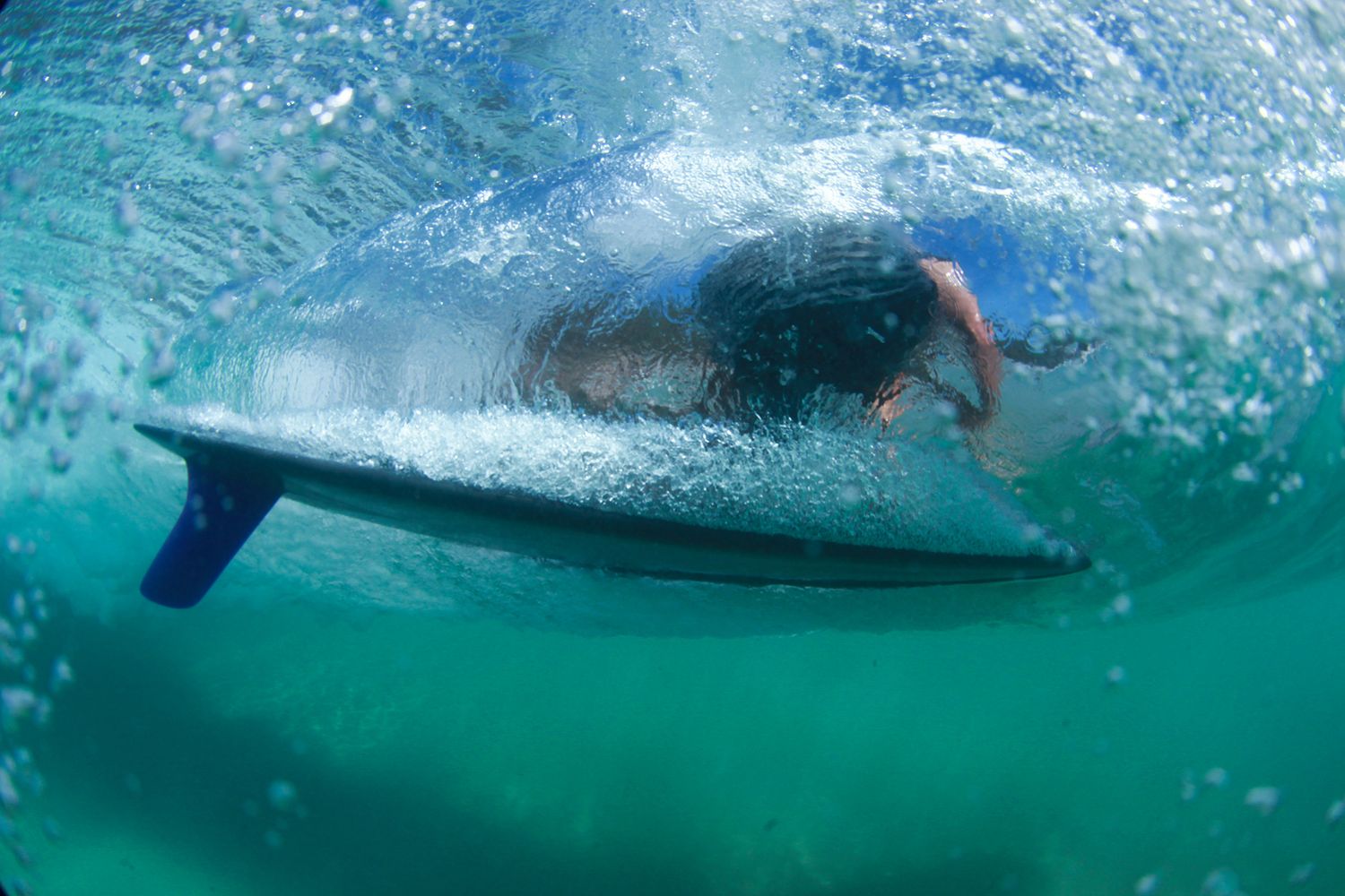 Underwater view of a surfer paddling through a wave on a surfboard in clear green ocean water.