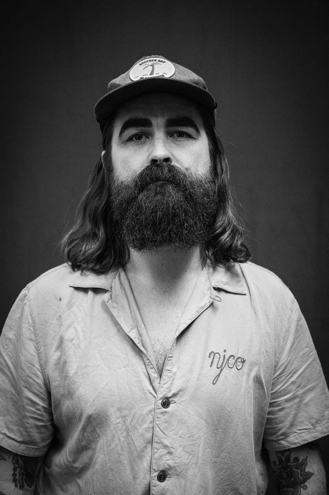 Black and white portrait of a bearded man with long hair wearing a cap and tattoos.