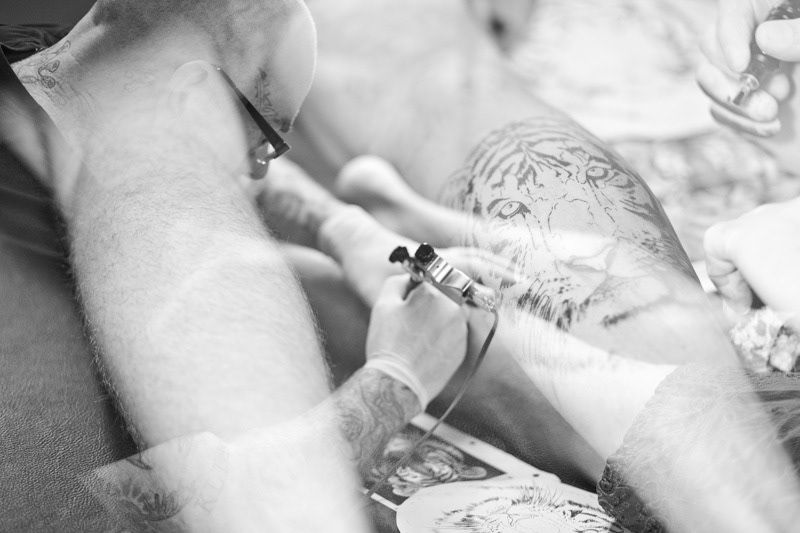 Black and white close-up of a tattoo artist applying detailed ink artwork to a clients leg.