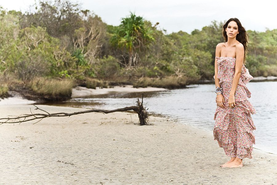 A woman in a flowing floral maxi dress stands barefoot on a sandy tropical beach.