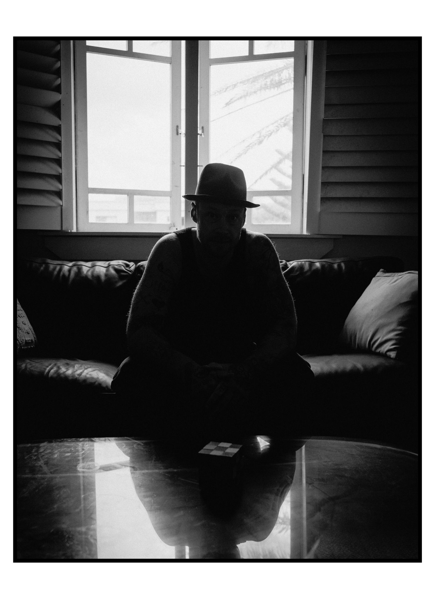 Silhouetted figure wearing fedora hat seated on leather sofa by bright window.