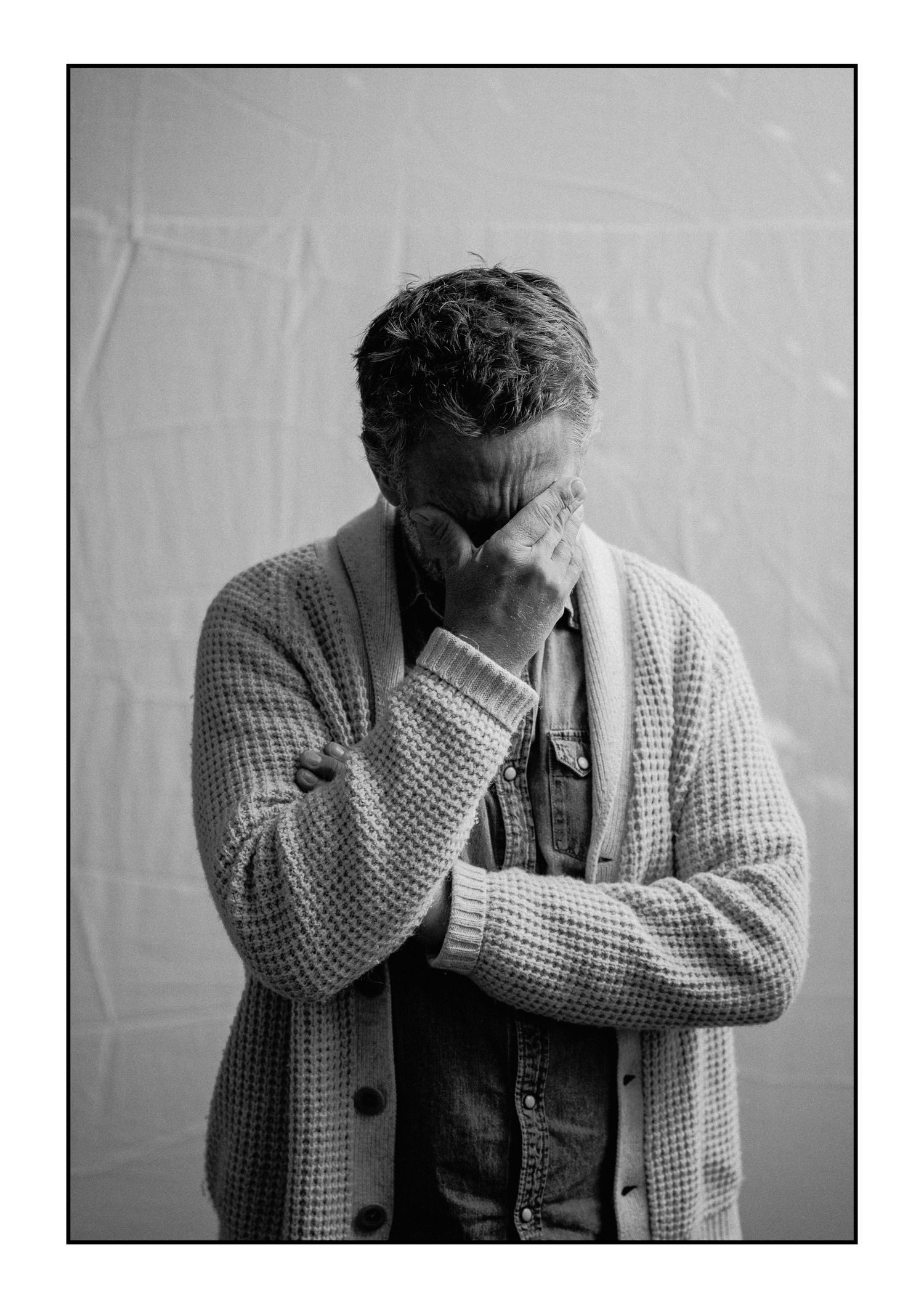 Man in knit cardigan bows head with hand covering face in emotional black and white portrait.