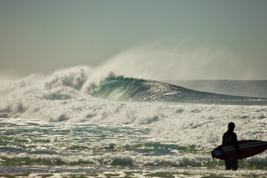 A lone surfer stands watching a massive powerful wave crashing on a dramatic coastline.