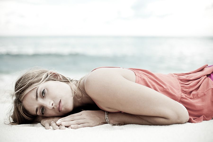 Young blonde woman in pink dress lying on white sand beach gazing at camera.