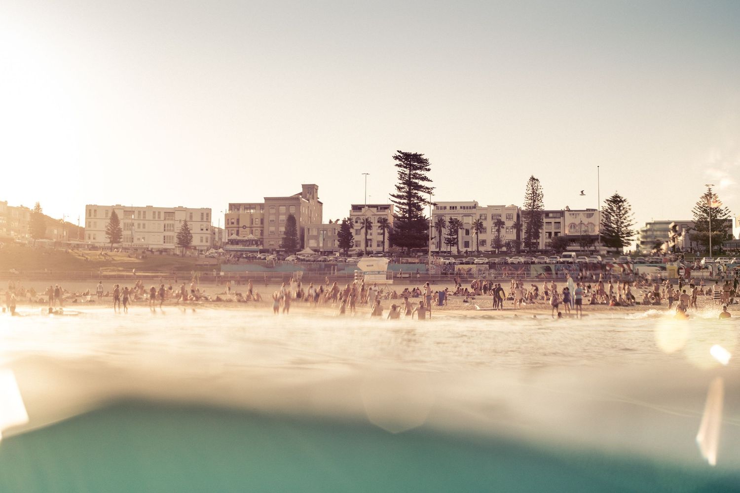 Sunny beach scene with crowds of people and buildings along the shoreline.