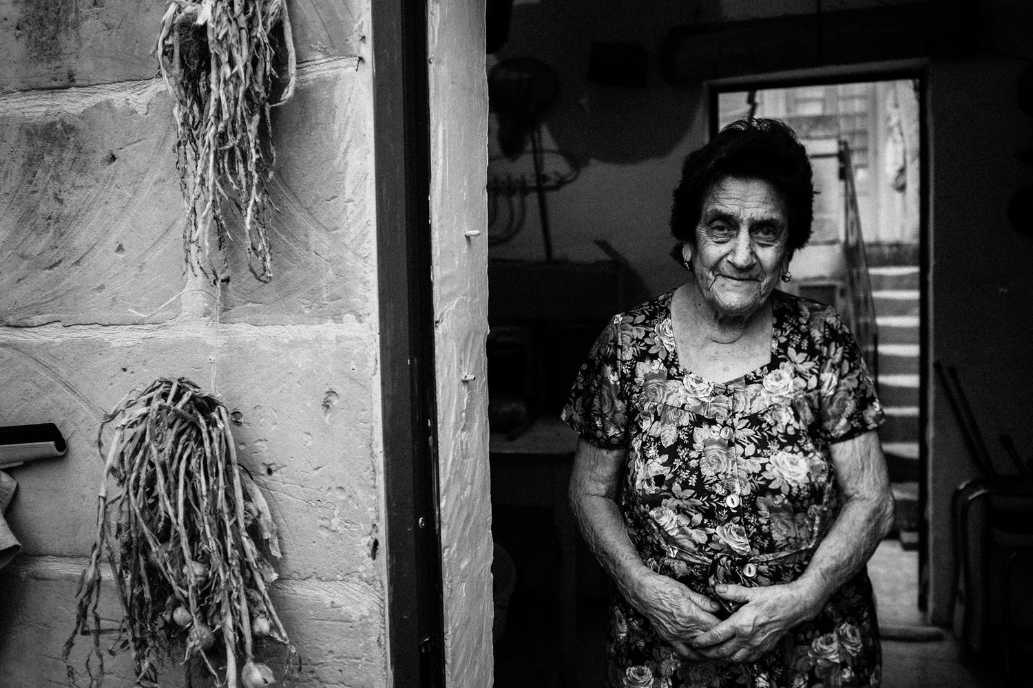 Black and white portrait of an elderly woman smiling in a doorway with dried plants.
