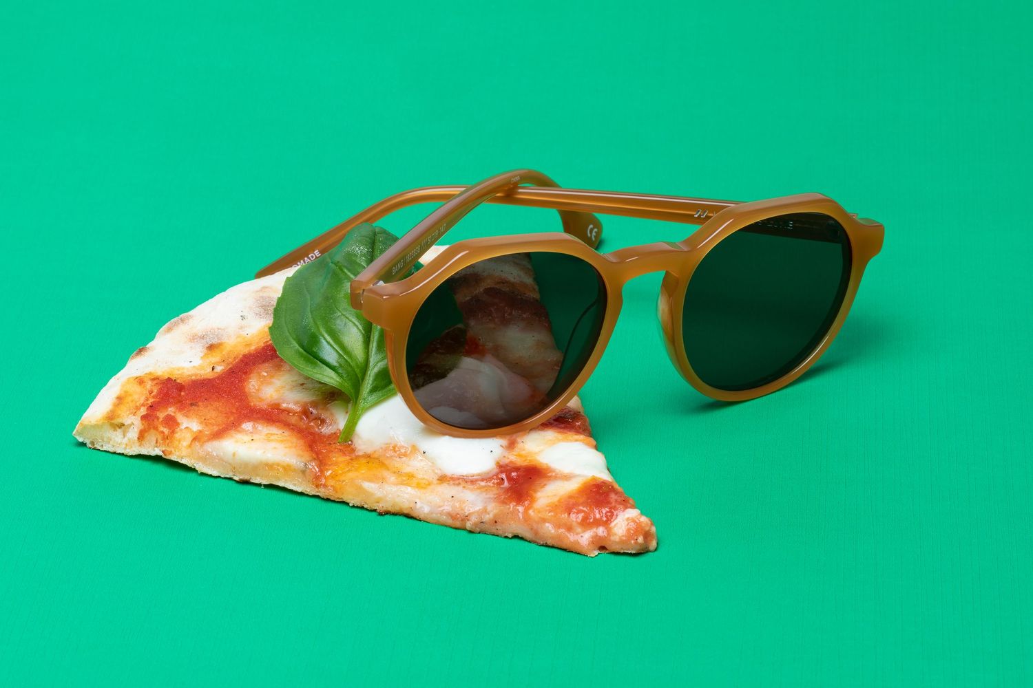 Trendy olive green round sunglasses resting on a fresh margherita pizza slice on green background.