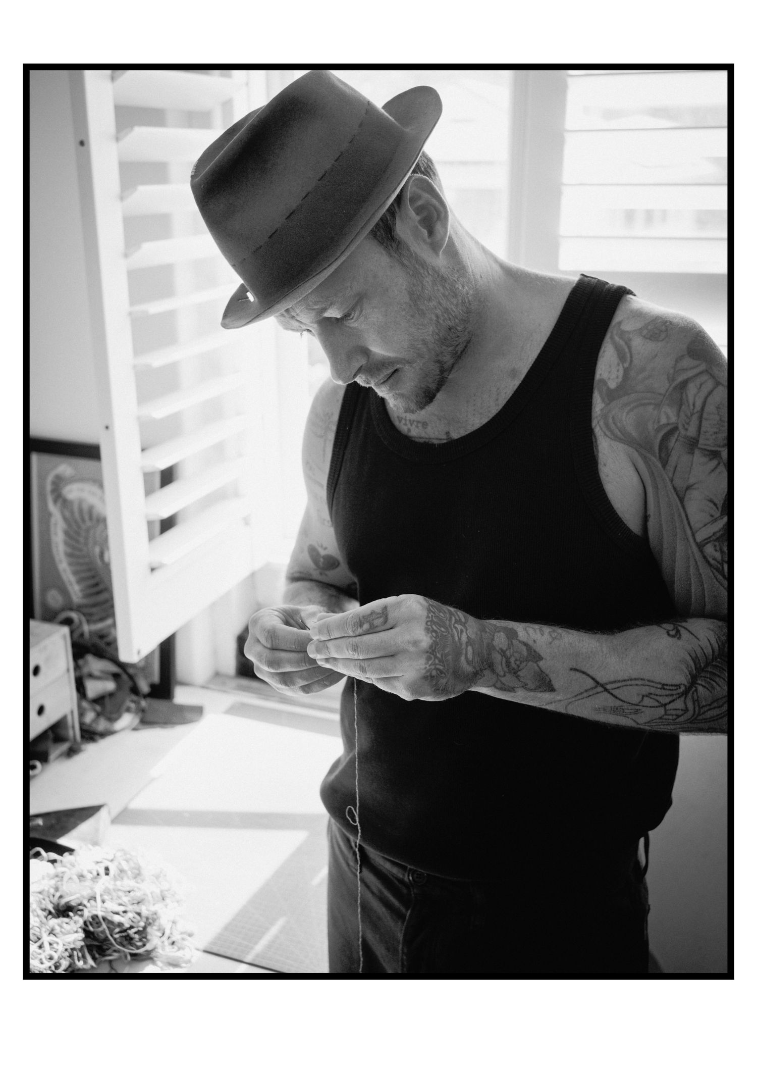 Tattooed man in black tank top and fedora examining small object in studio.