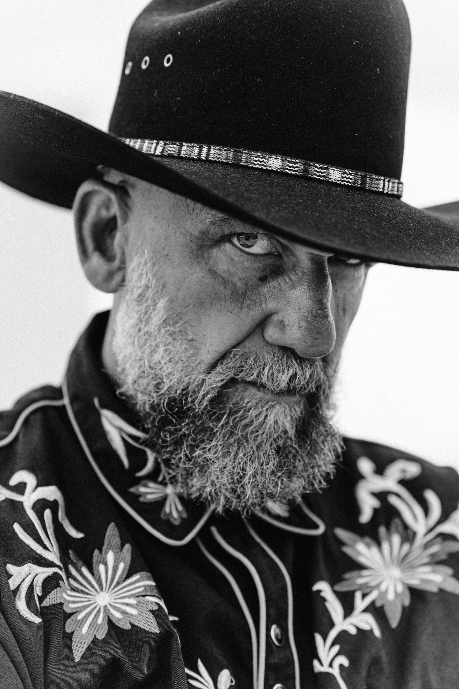 Dramatic black and white portrait of bearded man in cowboy hat and floral embroidered western shirt.