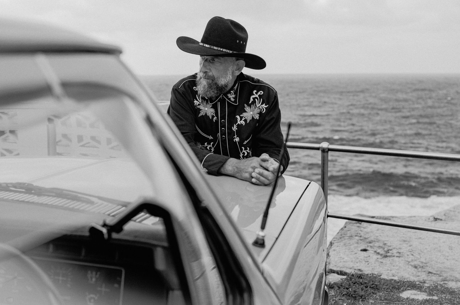 Man in cowboy hat leaning on vintage truck hood overlooking ocean, moody black and white photography.