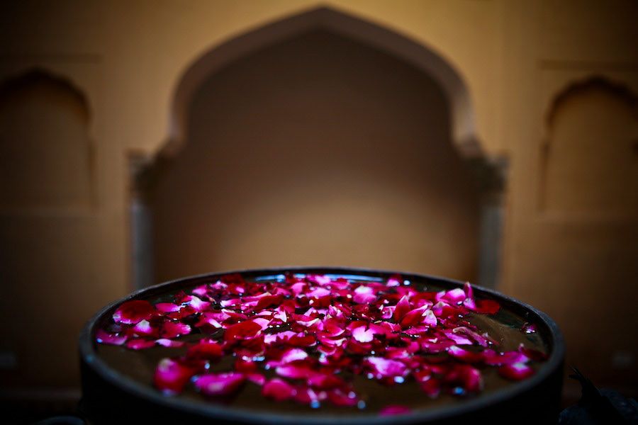Close-up of vibrant red rose petals floating in a dark bowl inside an ornate Indian palace interior.