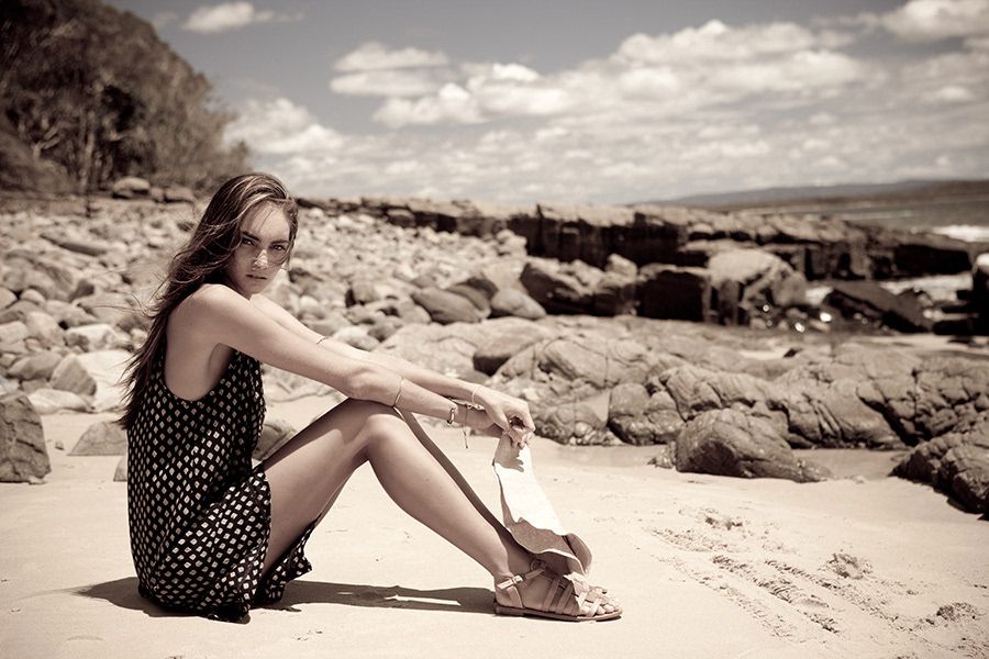 Woman in polka dot dress sitting on sandy beach with rocky coastline.