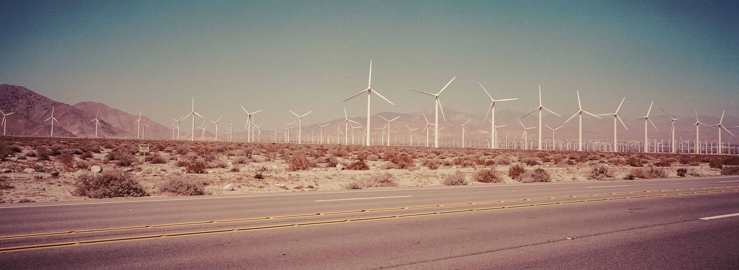 Panoramic view of a large wind farm with white turbines along a desert highway with mountain backdrop.