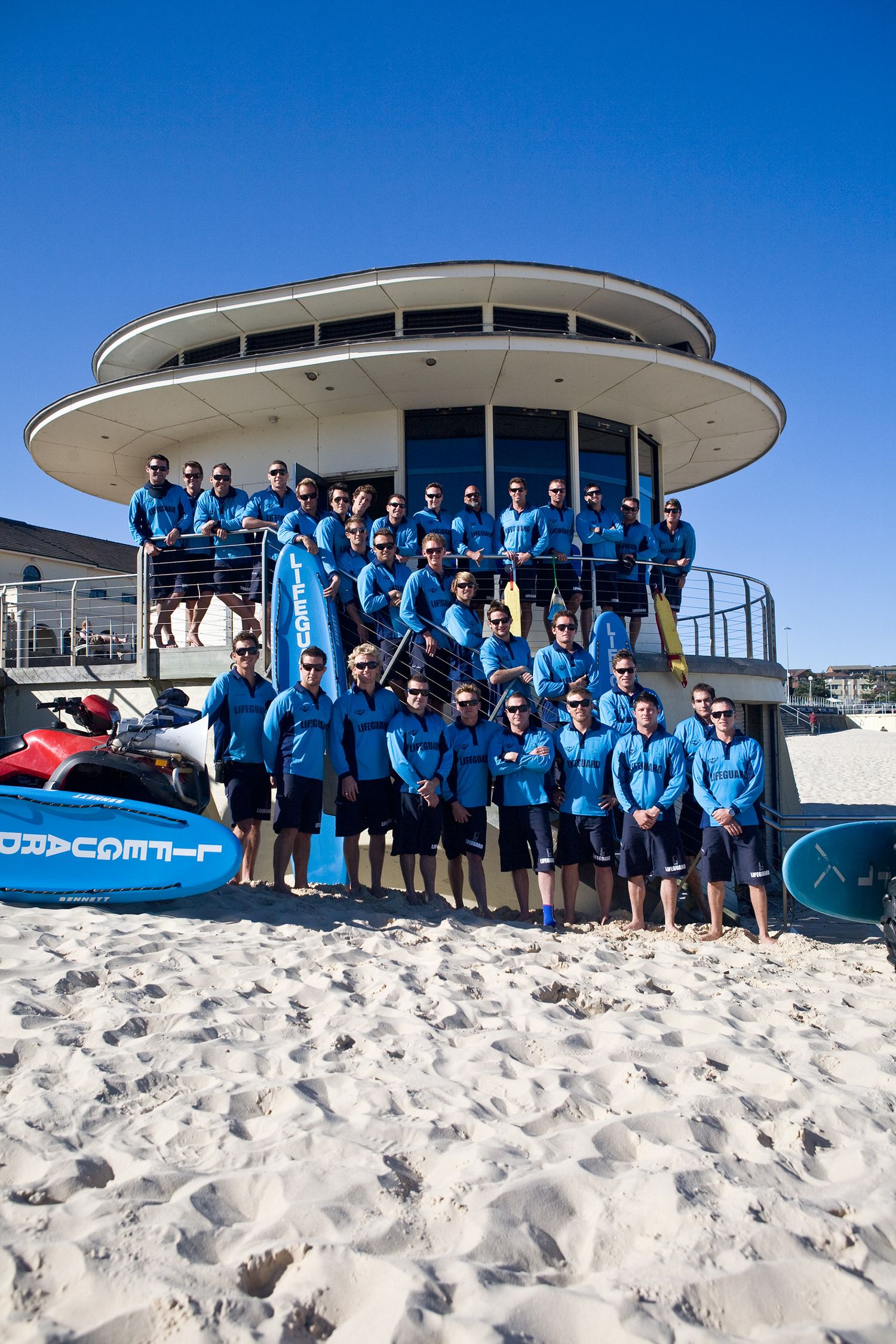 Large group of lifeguards in matching blue uniforms posing outside a beach patrol building.