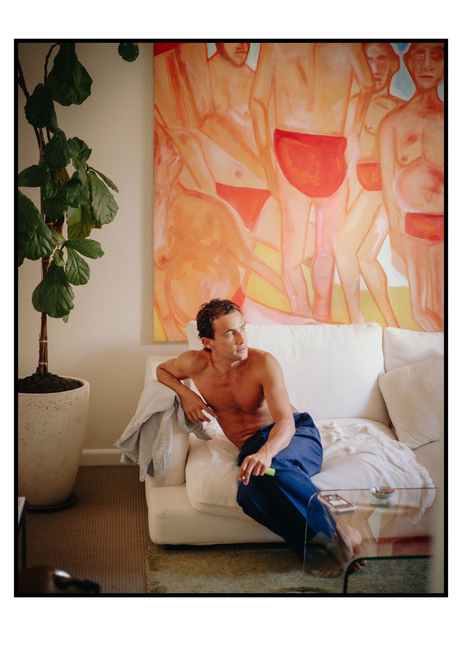Shirtless man in blue pants relaxes on white sofa beneath large colorful figurative painting indoors.