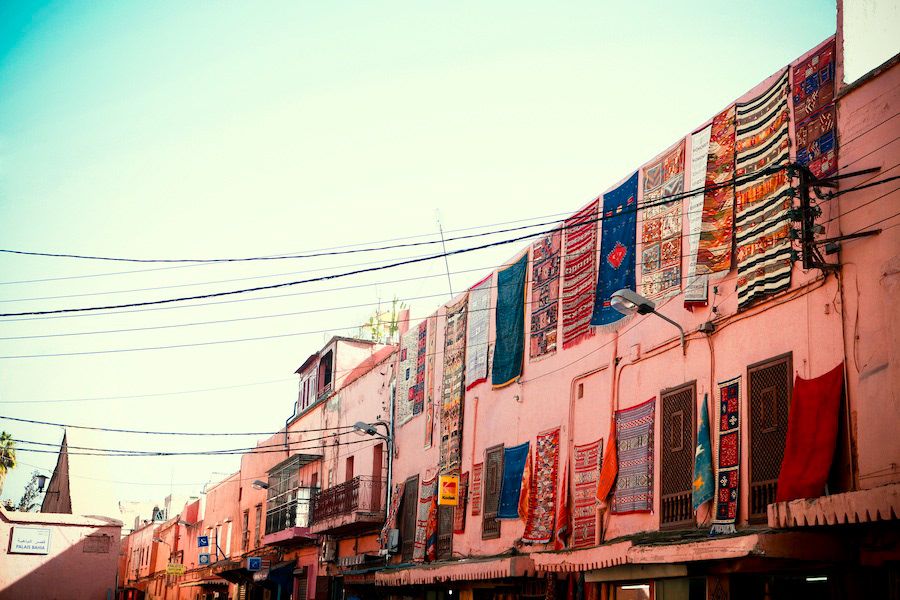 Colorful textiles and rugs hang from pink building facades lining a vibrant Moroccan market street.