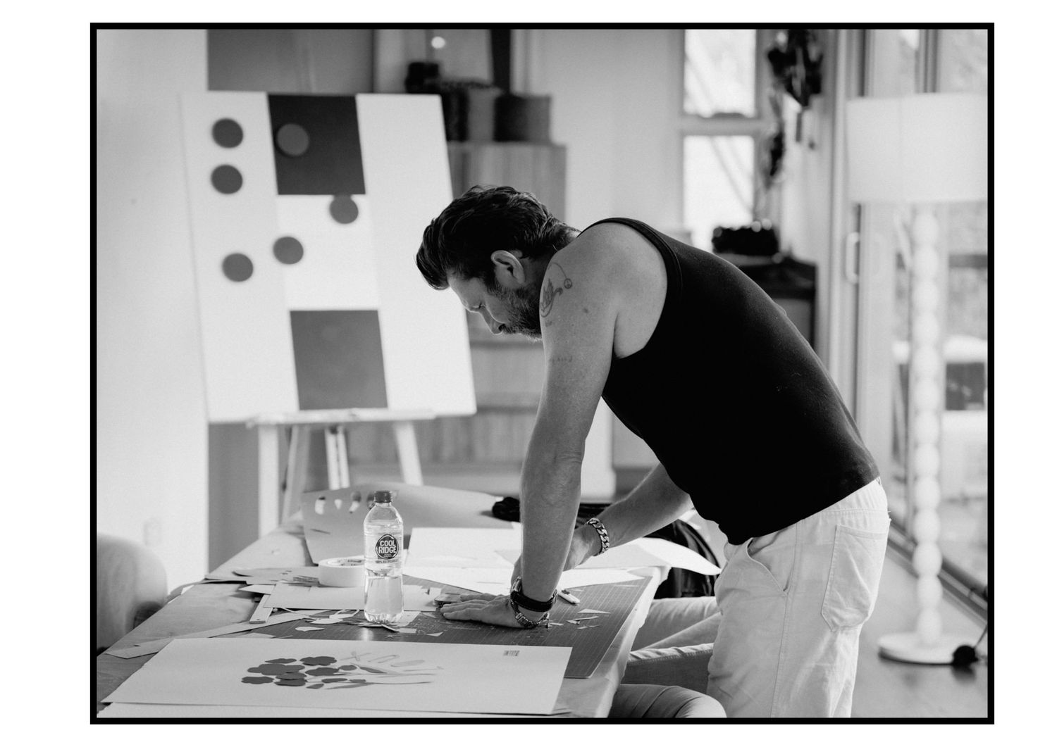 Artist in black tank top leaning over worktable in studio in black and white.