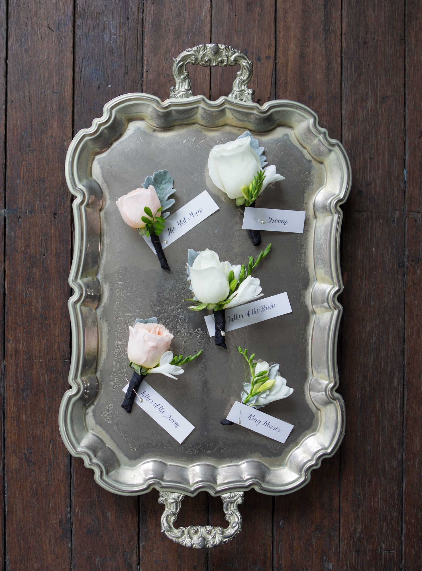 White and blush rose boutonnieres with name tags arranged on an ornate silver serving tray.