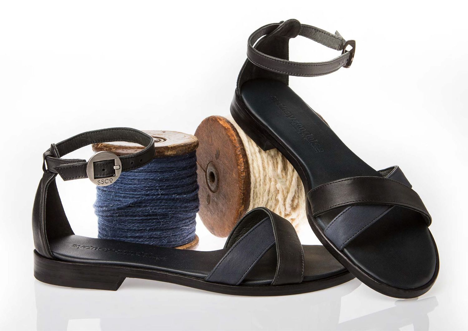 Black leather flat sandals with ankle strap displayed next to wooden thread spools on white background.