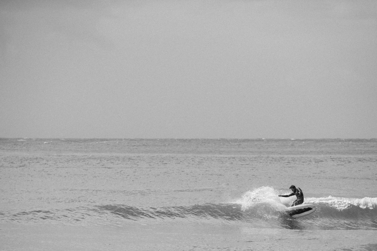 Black and white photo of a lone surfer riding a small wave on a calm ocean.
