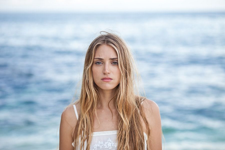 A young blonde woman in a white top stands before a blue ocean with windswept hair.