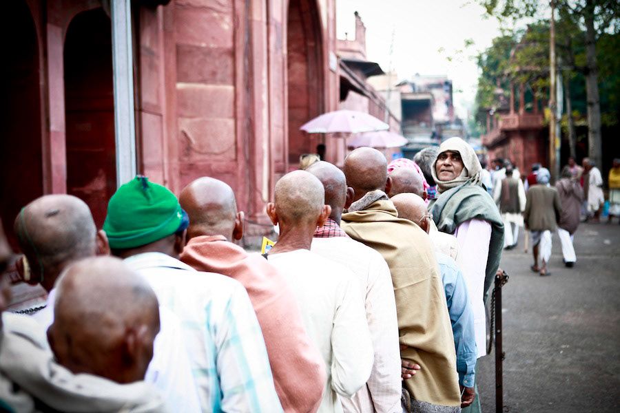 Line of shaved-head Hindu pilgrims queuing outside red sandstone temple in India.
