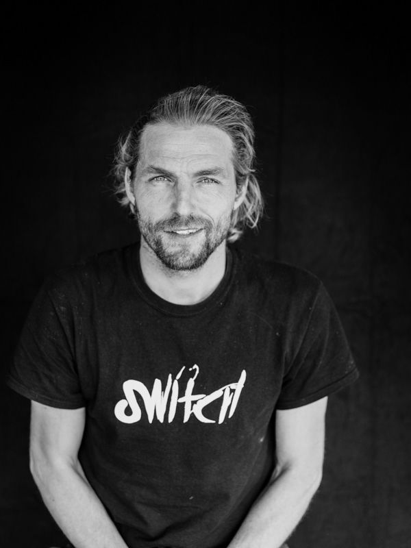 Black and white portrait of a smiling bearded man wearing a Switch brand graphic t-shirt.