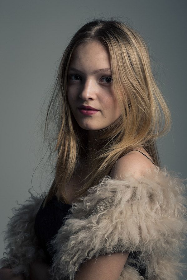 Studio portrait of a young blonde woman wearing a fluffy fur coat against a gray background.