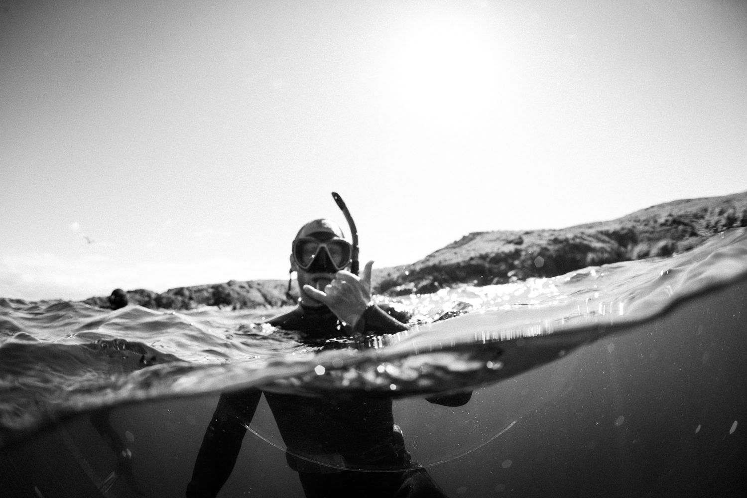 Black and white split-level photo of snorkeler at ocean surface with rocky coastline behind.