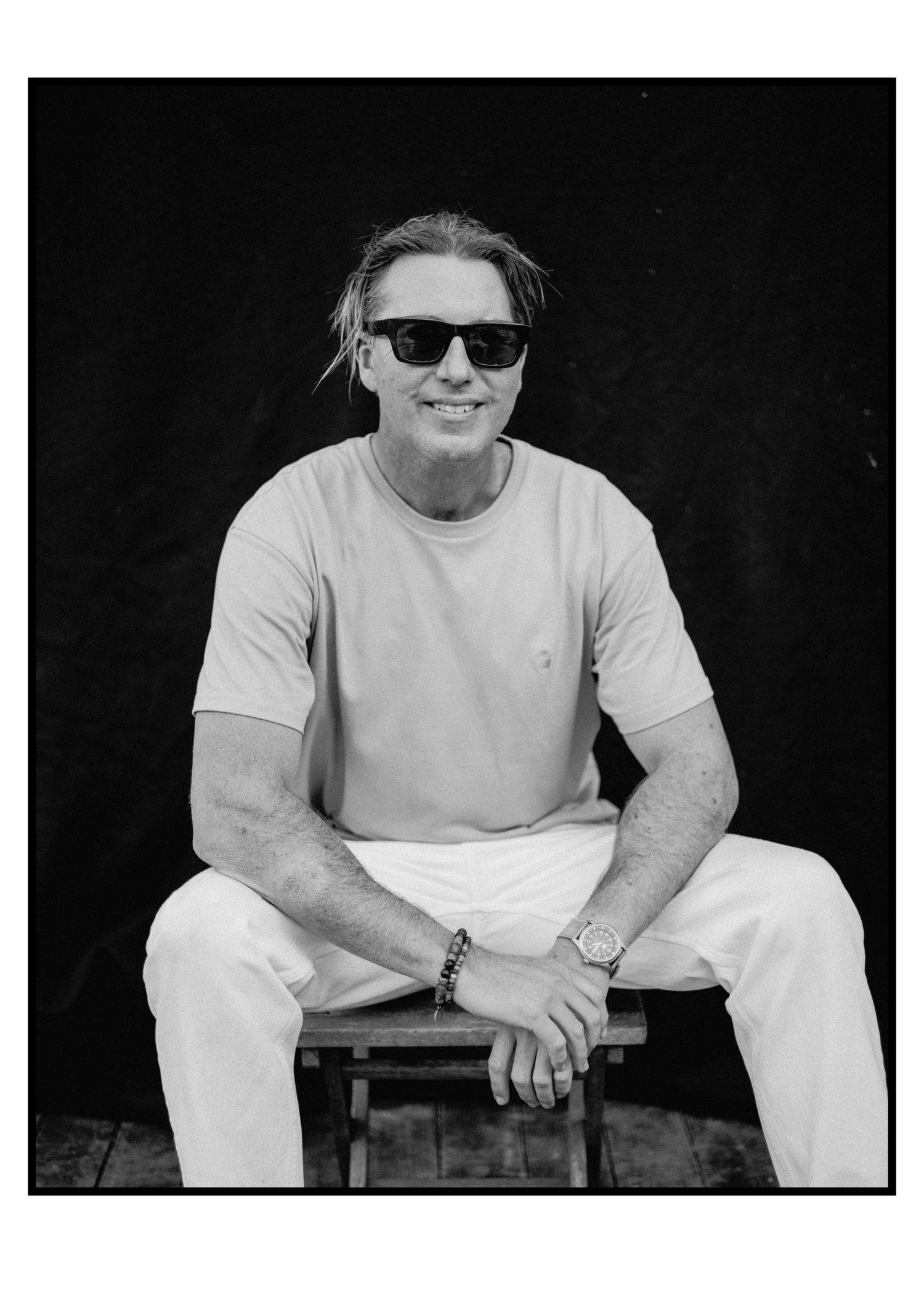 Smiling man in sunglasses and light casual outfit sits relaxed on stool in black and white portrait.