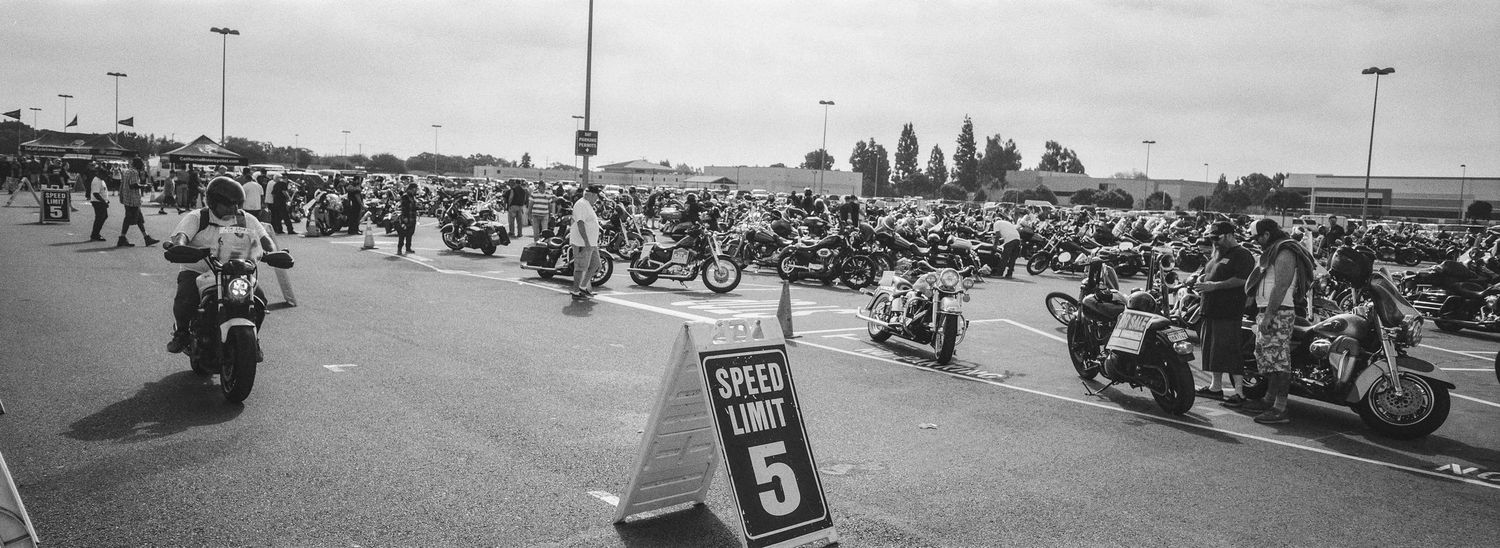 Black and white panoramic view of large motorcycle rally in parking lot with speed limit sign.