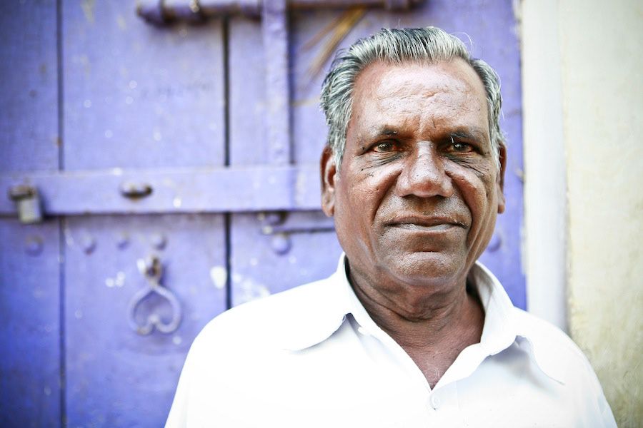 Portrait of a mature Indian man in a white shirt standing before a rustic blue wooden door.